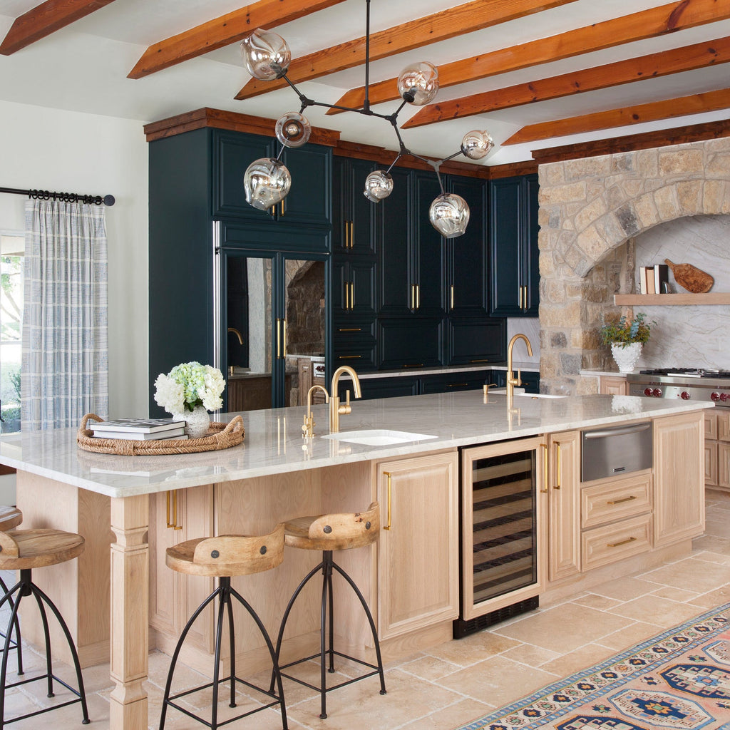 Clear globes on dark arms form this Clérachic chandelier, hanging below orange beams above a kitchen island with a stone arch behind.
