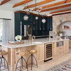 Clear globes on dark arms form this Clérachic chandelier, hanging below orange beams above a kitchen island with a stone arch behind.