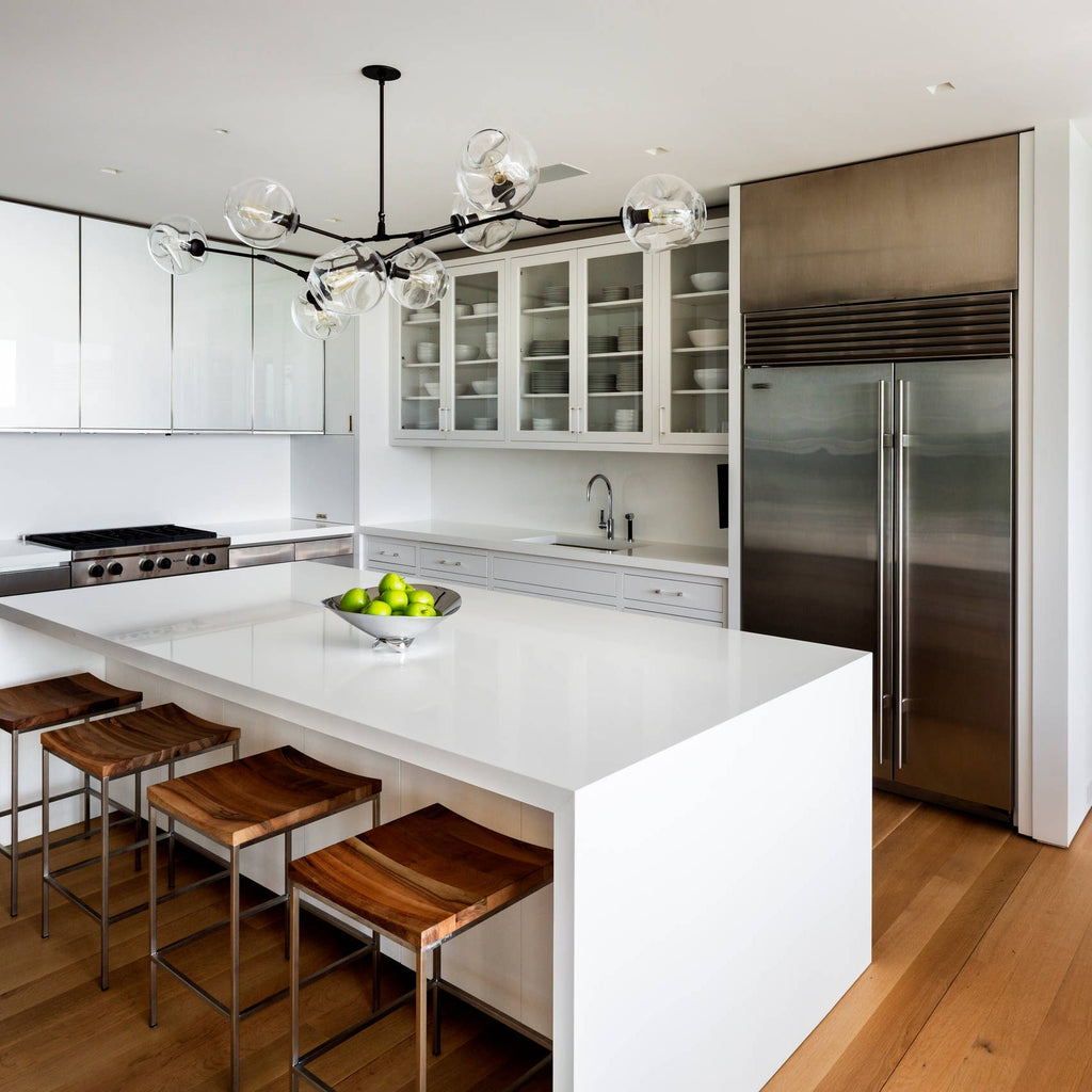 This Clérachic chandelier with clear globes hangs above a white waterfall kitchen island, with a steel fridge and wooden stools nearby.