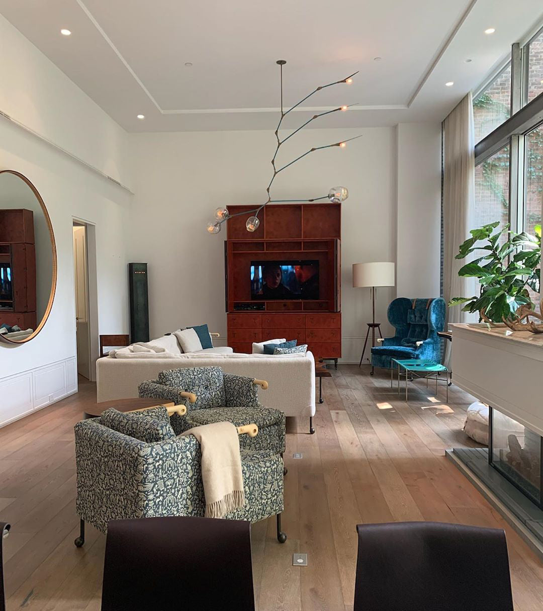 A tall Clérachic chandelier with thin branching arms hangs in a high-ceilinged living room with a round mirror and teal armchair.