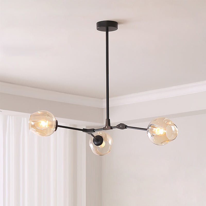 Black-framed Clérachic chandelier with three amber glass globes hanging from a short rod in a pale room with white curtains.