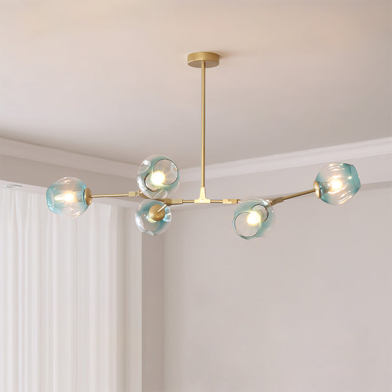 Gold Clérachic chandelier with five gradient blue glass globes lit softly, hung from a slim rod in a pale room with white curtains.