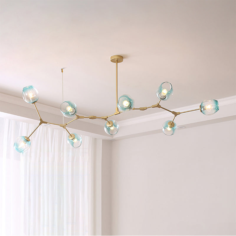 Long gold Clérachic chandelier with nine gradient blue glass globes lit along a branching frame, hung in a pale corner room.
