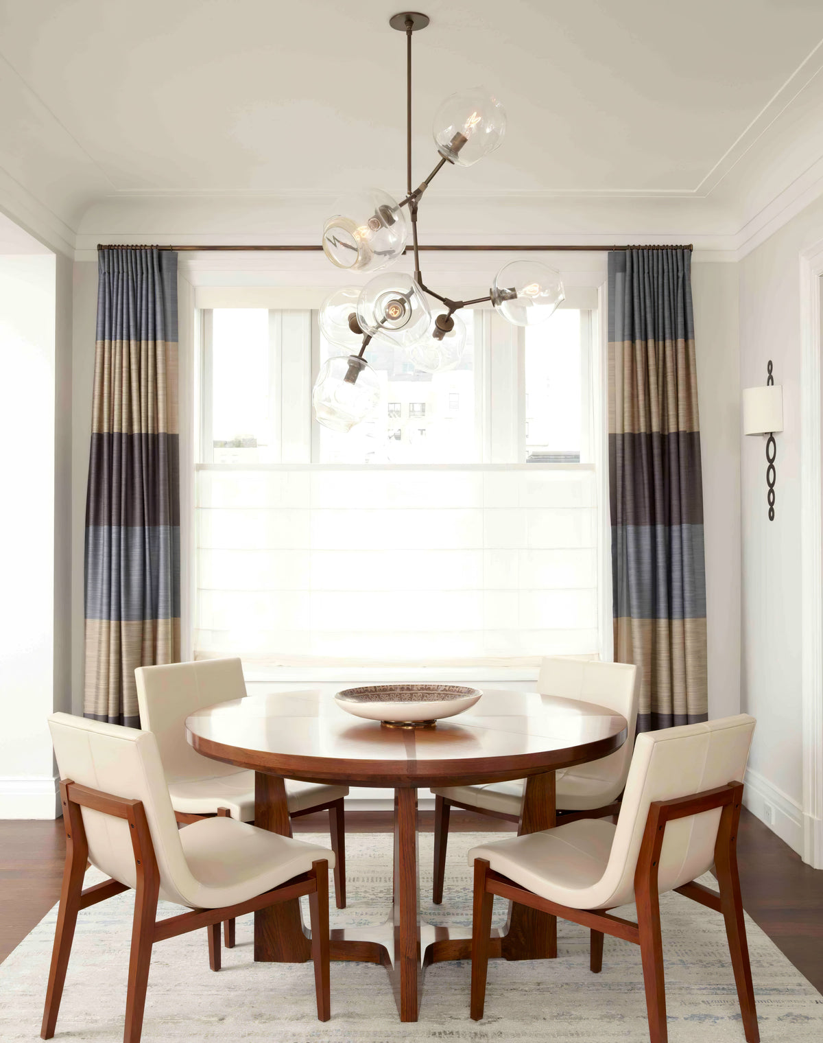 A Clérachic chandelier above a round wooden dining table with cream chairs, in front of striped curtains.