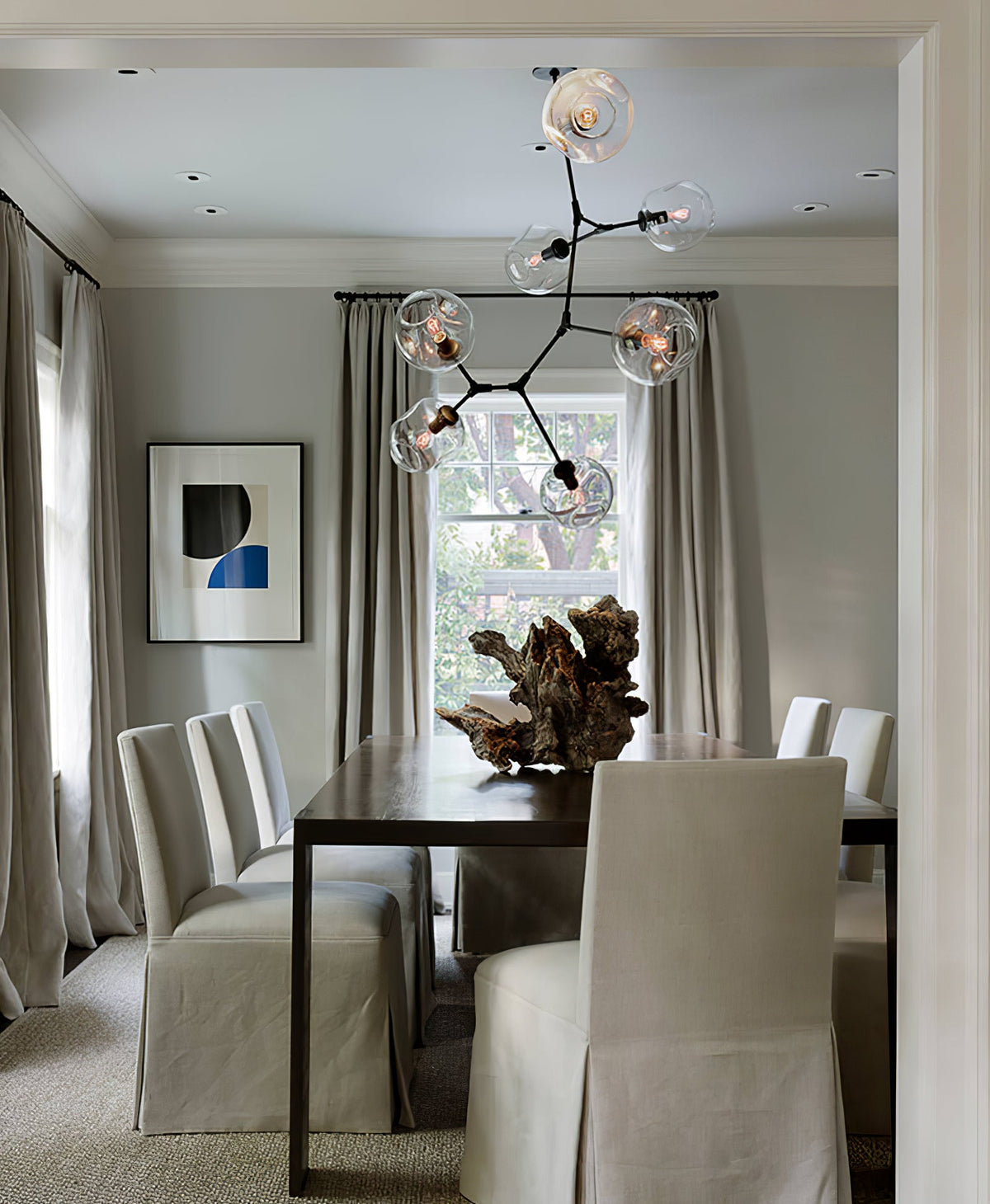 Clérachic chandelier with clear globes above a dark dining table with grey slipcovered chairs, in front of a sash window.