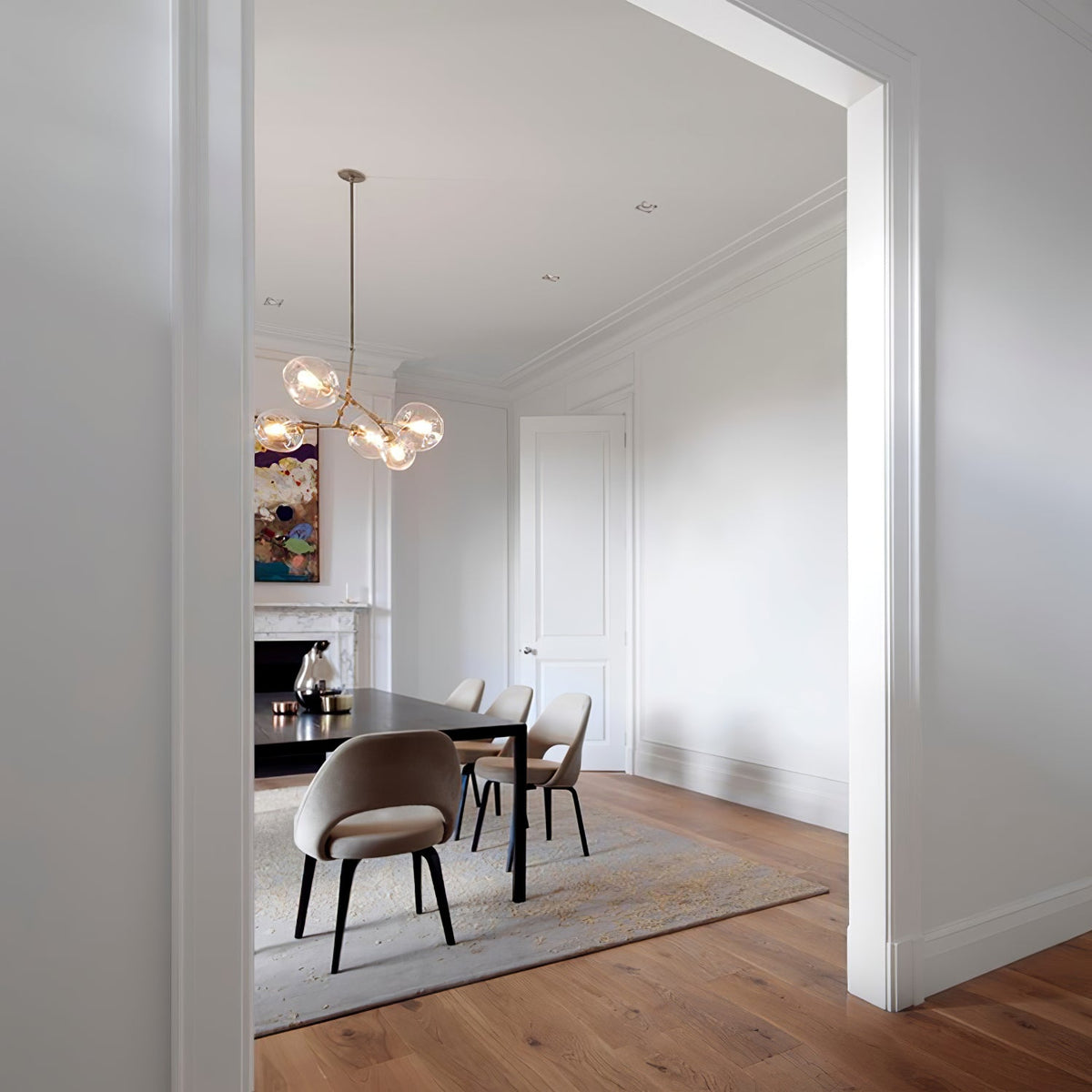 A Clérachic chandelier with brass arms hanging in a pale dining room with a dark table, upholstered chairs and wood flooring.