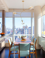 Clérachic chandelier with brass arms and a few clear globes in front of a floor-to-ceiling city window, over a dining table.
