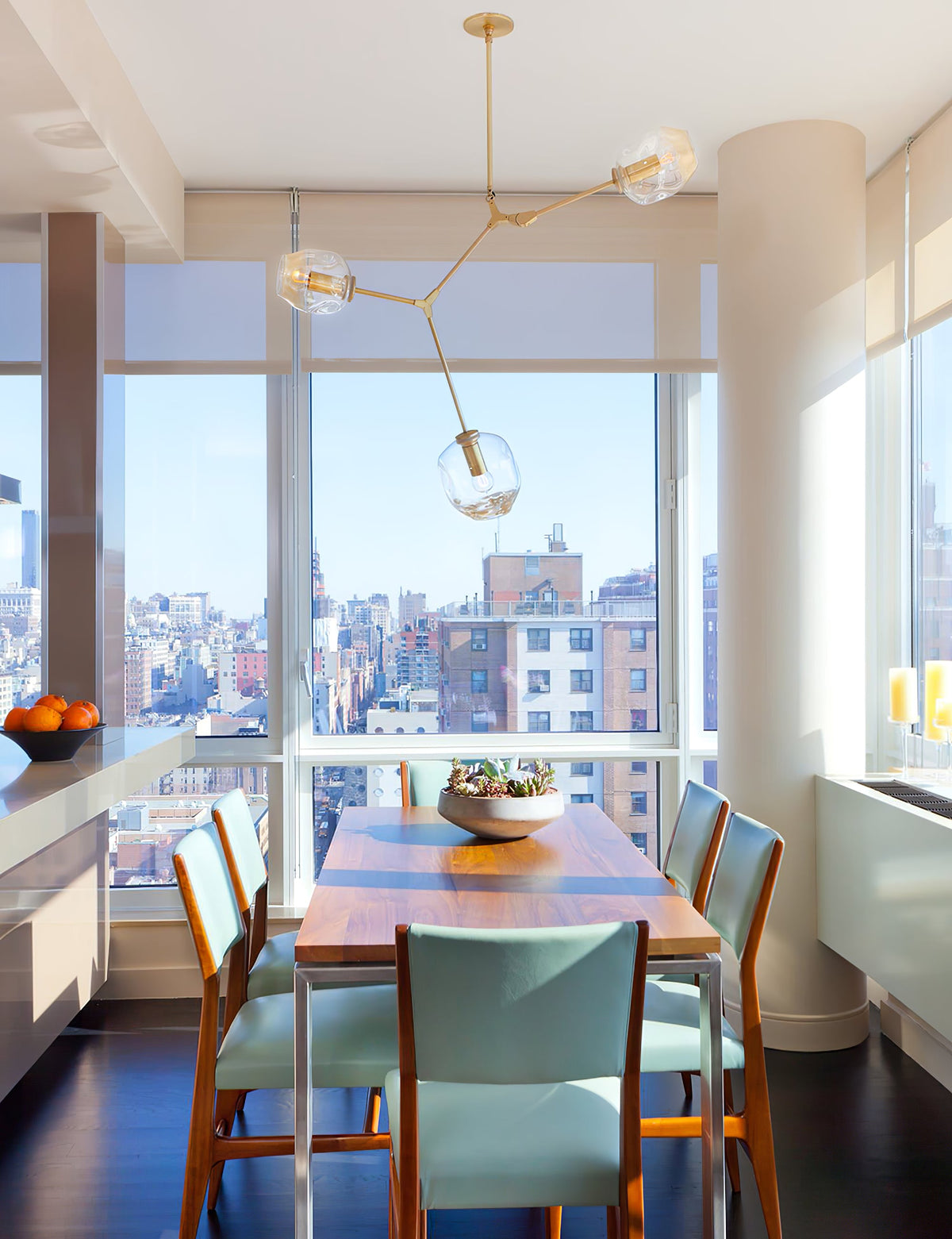 Clérachic chandelier with brass arms and a few clear globes in front of a floor-to-ceiling city window, over a dining table.