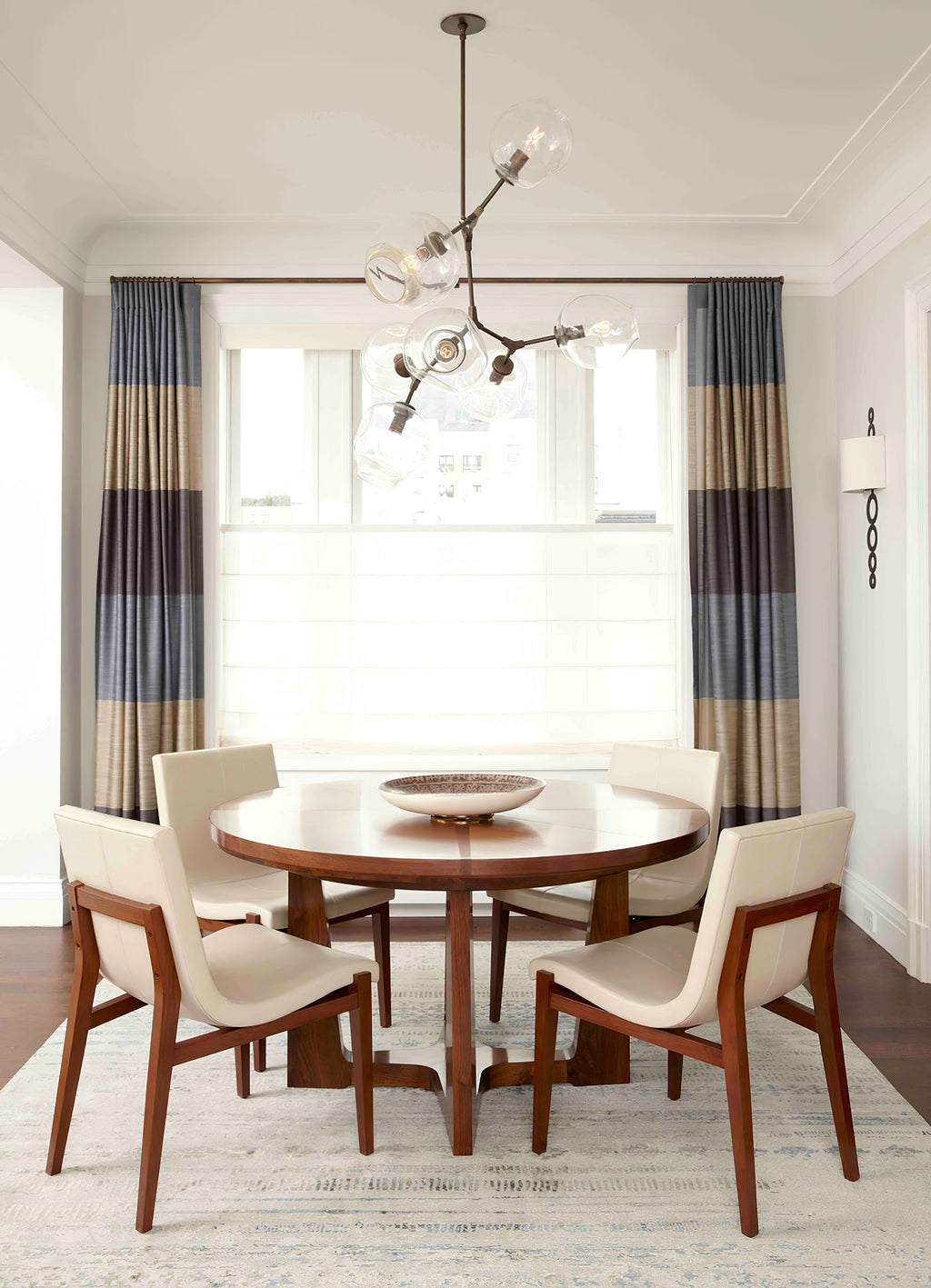 Clérachic chandelier with clear globes above a round dining table and cream chairs, framed by blue and grey striped curtains.