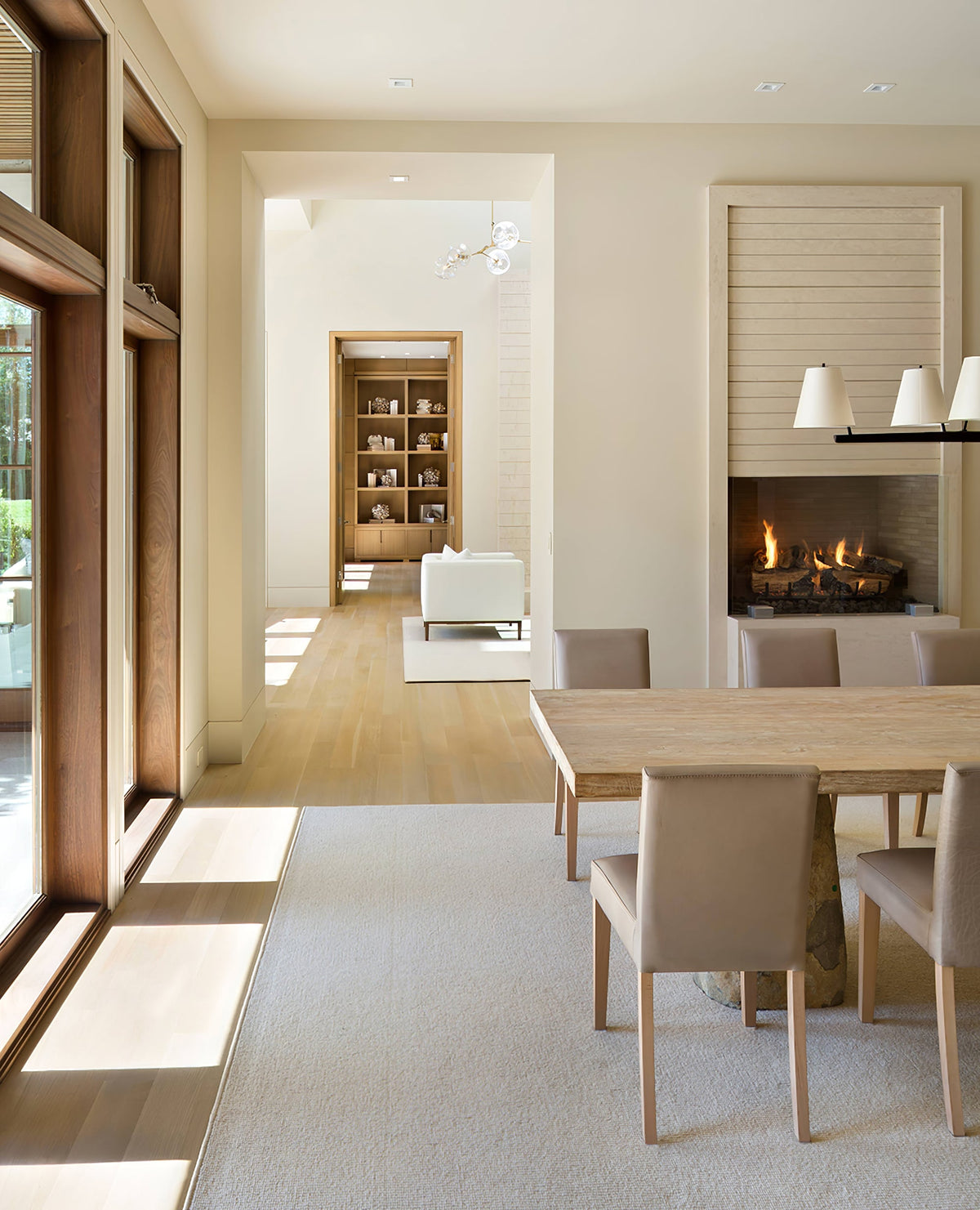A Clérachic chandelier seen far down a long, bright room with a dining table, a fireplace and tall wood-framed windows.