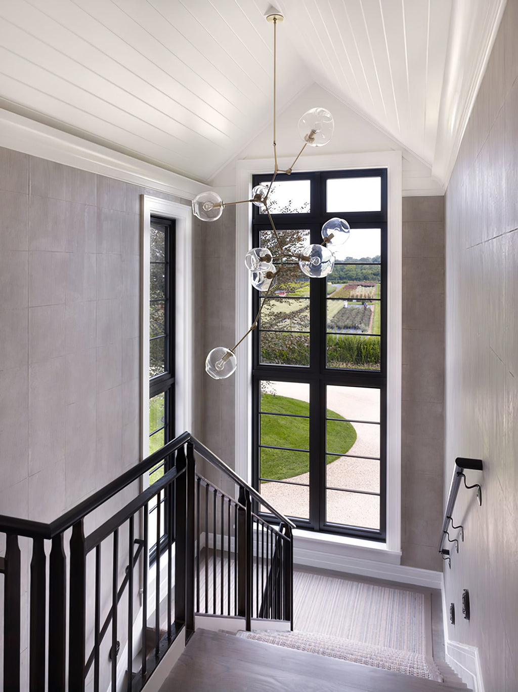 Clérachic chandelier with clear globes hanging through a stairwell beside a tall black-framed window and a black balustrade.