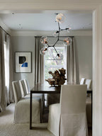 Clérachic chandelier with clear globes above a dark dining table with slipcovered chairs, a driftwood piece and a window behind.