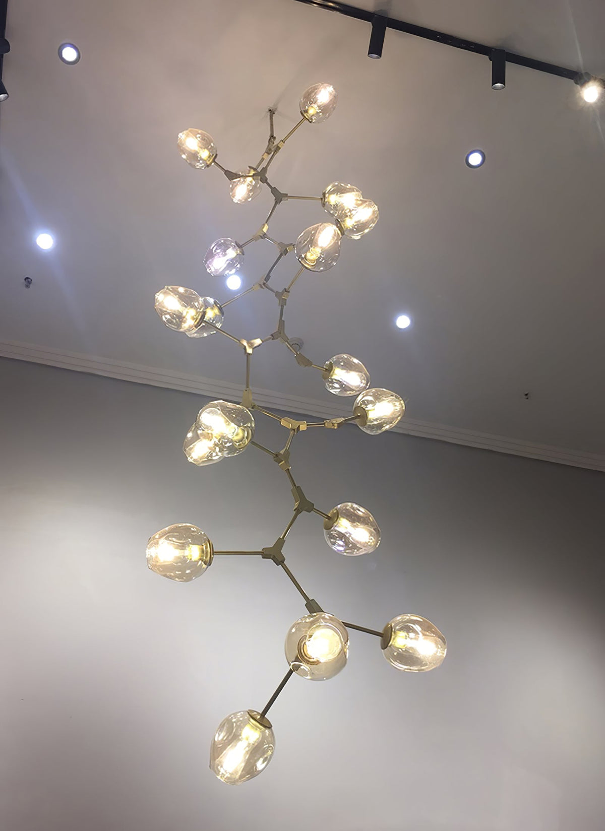 Looking up at a lit Clérachic chandelier with brass arms and clear globes, hanging long under a high ceiling with track lights.
