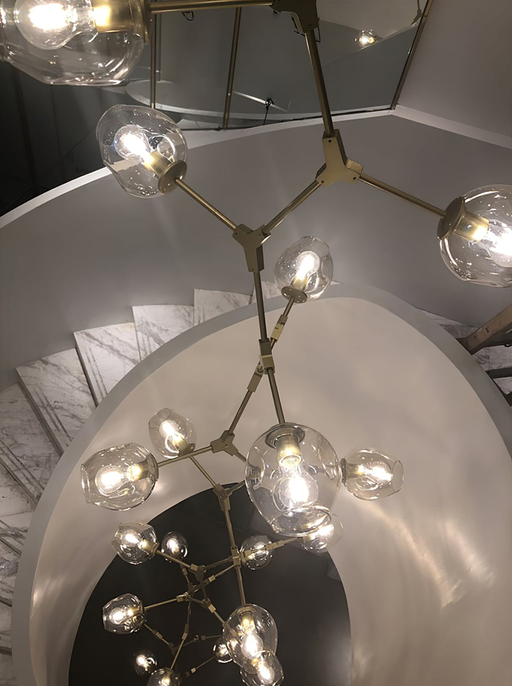 Looking down a spiral stair, with a lit Clérachic chandelier in brass and clear globes branching out in the foreground.