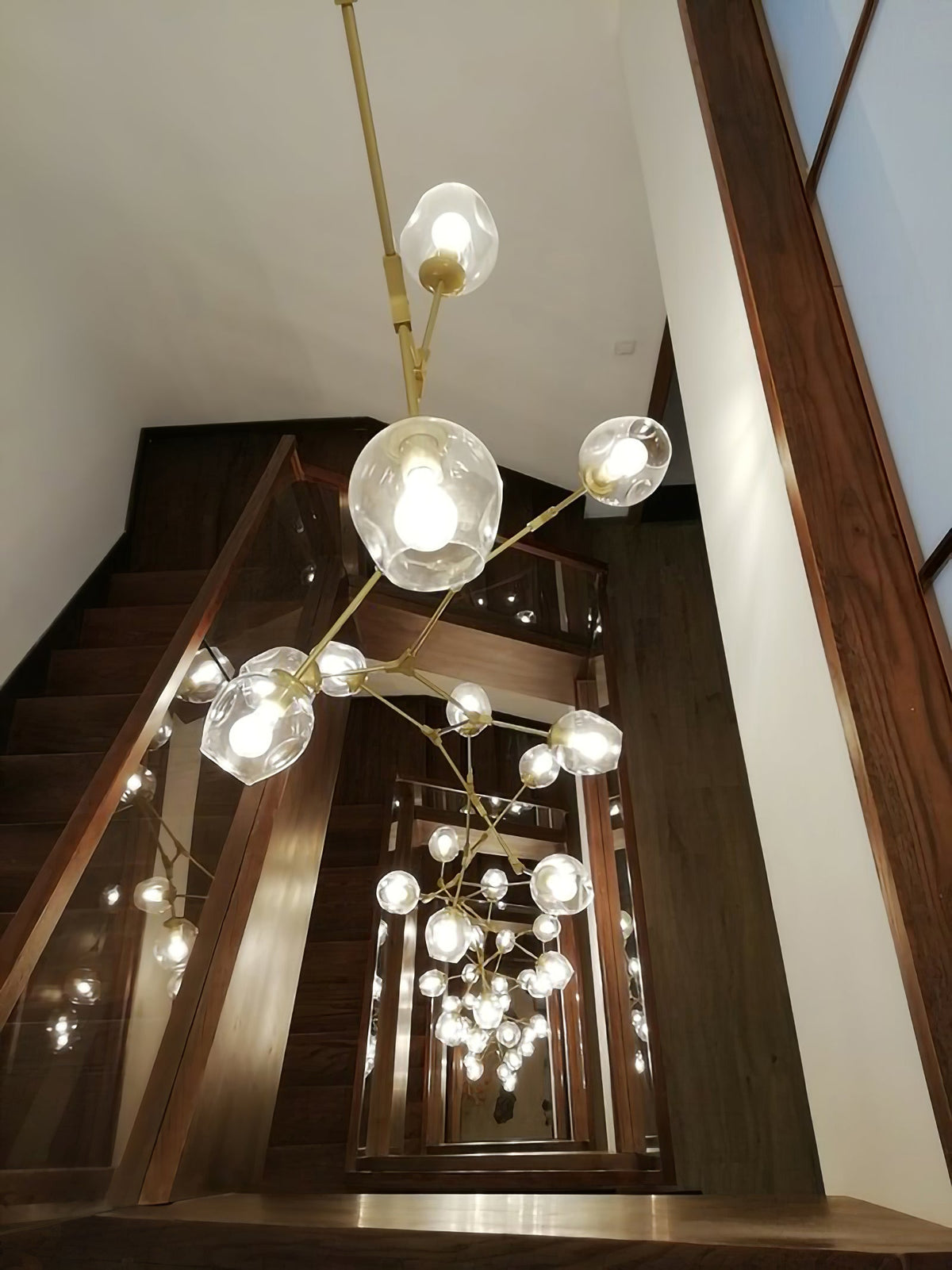 Looking up a timber stairwell at a lit Clérachic chandelier with brass arms and clear globes, repeated by the glass beyond.