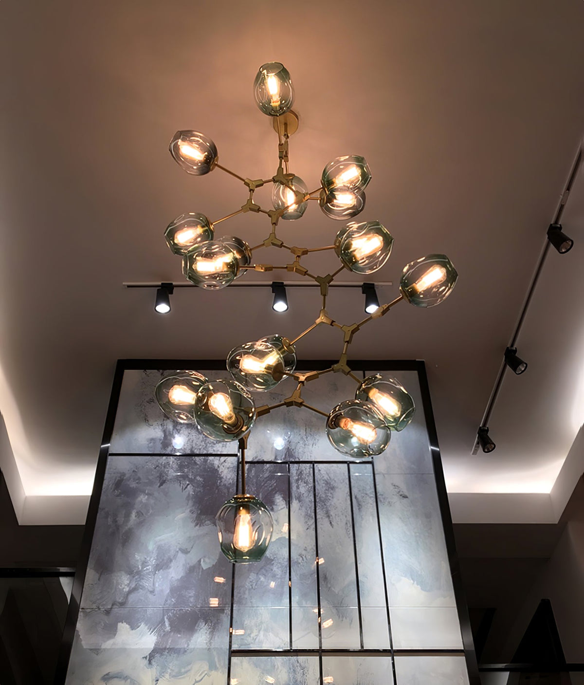 Lit Clérachic chandelier in brass with clear globes hanging in a double-height space above track lights and a glazed wall.