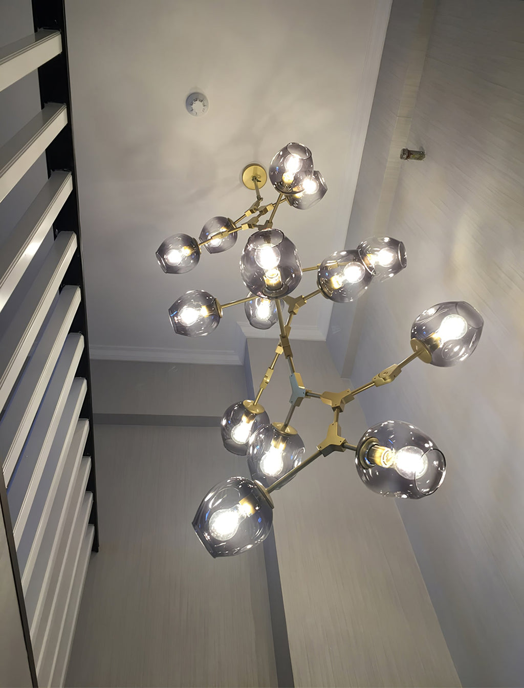 Looking up at a lit Clérachic chandelier with brass arms and smoky grey globes in a narrow stairwell.