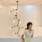 A woman stands in a curved white room beside a Clérachic chandelier with black arms and warm-lit globes on a long ceiling rod.
