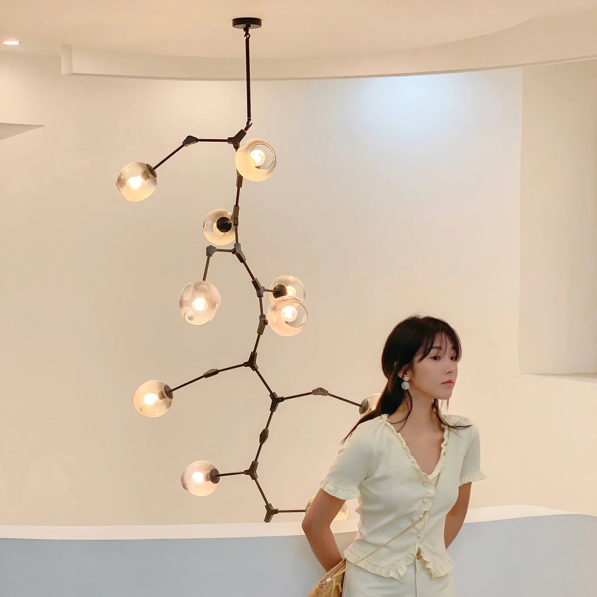 A woman stands in a curved white room beside a Clérachic chandelier with black arms and warm-lit globes on a long ceiling rod.