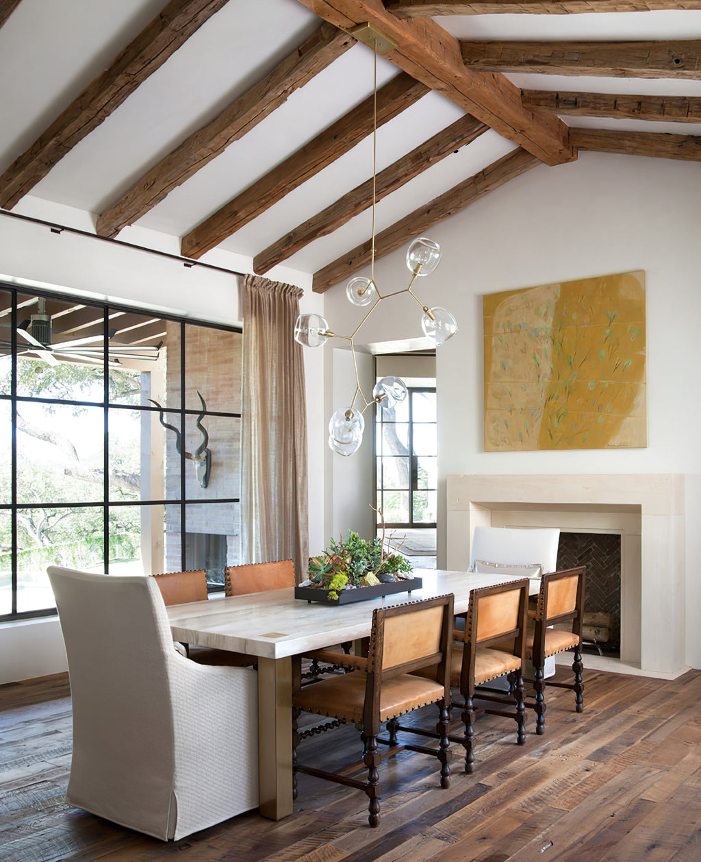 Clérachic chandelier with clear globes in a dining room with exposed timber roof beams, a limestone table and a fireplace.