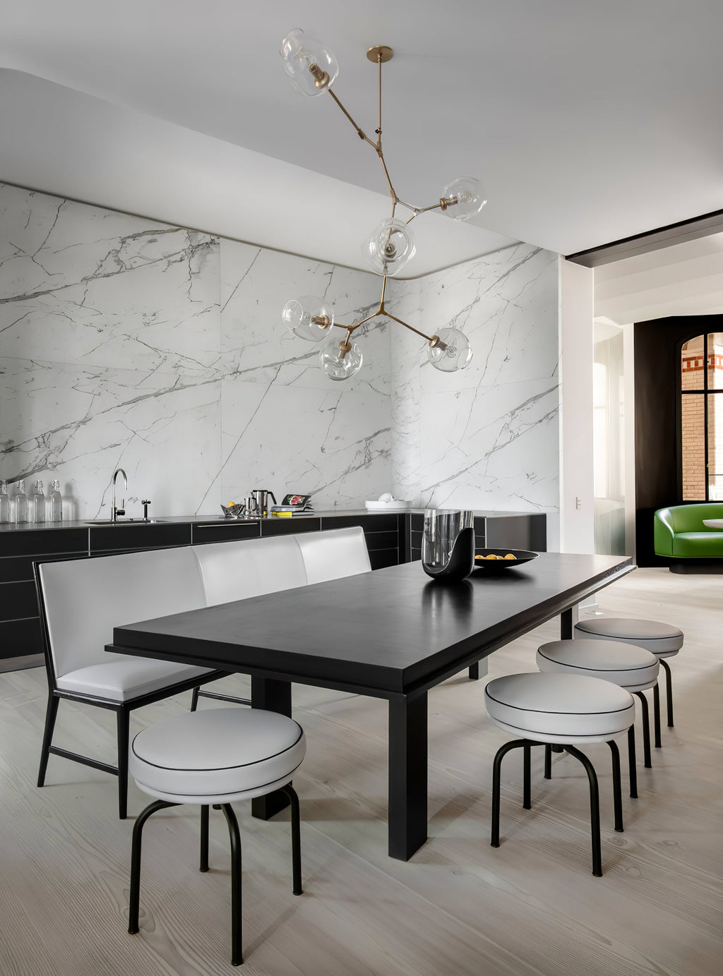 Clérachic chandelier with brass arms and clear globes over a long black table with stools, in front of a marble wall.
