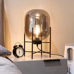 Lit amber glass shade glowing warmly on this Clérachic table lamp, beside an alarm clock on a grey bedside cabinet.