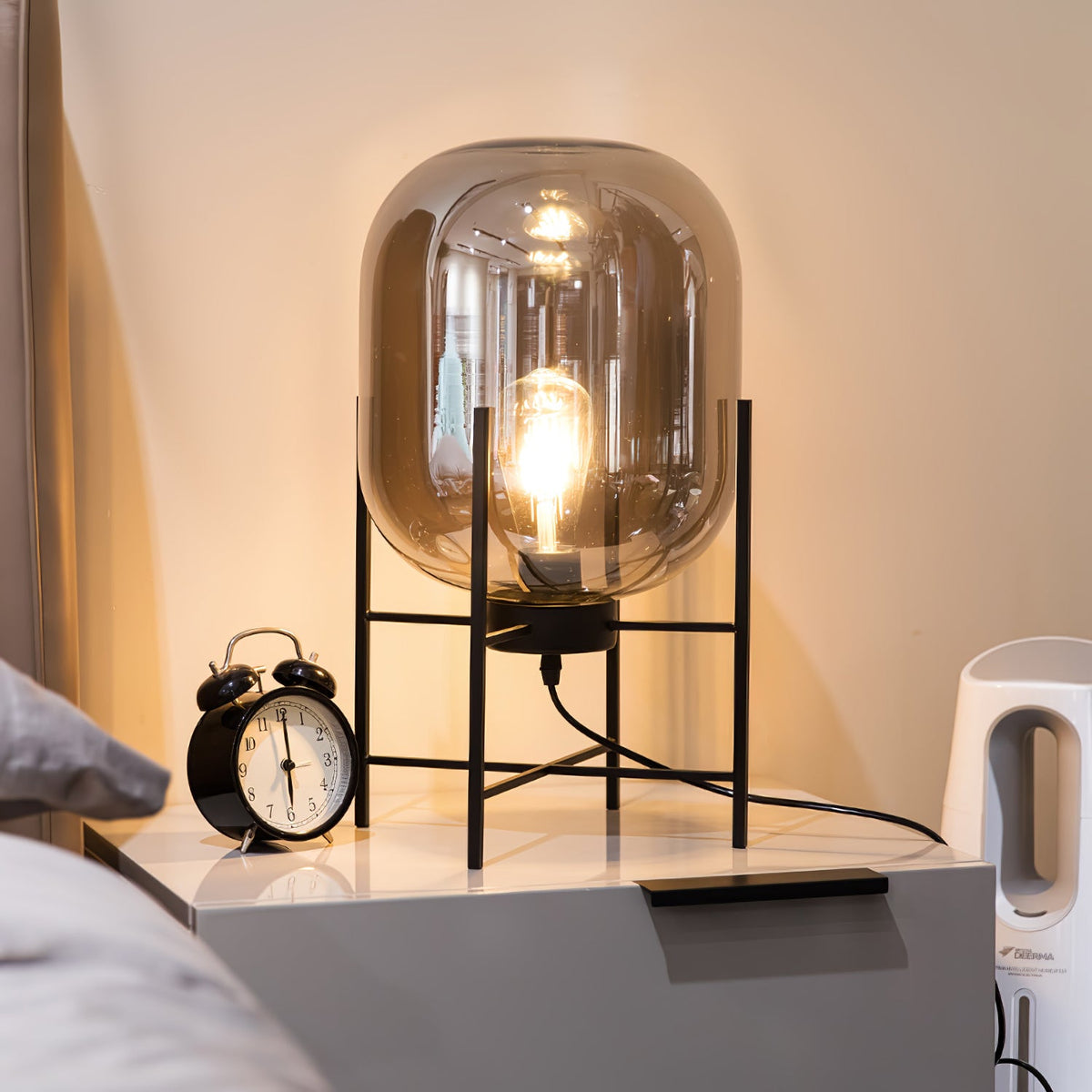Lit amber glass shade glowing warmly on this Clérachic table lamp, beside an alarm clock on a grey bedside cabinet.