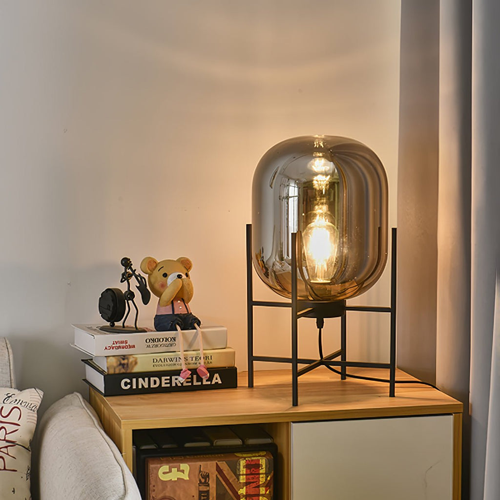Smoke-grey glass shade lit from within on a black stand, this Clérachic lamp sitting on a pale wooden nightstand with books.