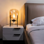 This Clérachic table lamp lights a bedside cabinet with a warm amber glow, next to a leather headboard and grey bedding.