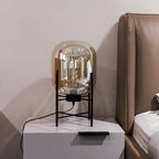 Close-up of the champagne-toned glass shade and black four-leg stand of this Clérachic lamp on a grey bedside unit.
