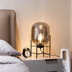 A dim, amber-lit glass shade on a black frame beside a bed with an alarm clock, from this Clérachic table lamp range.