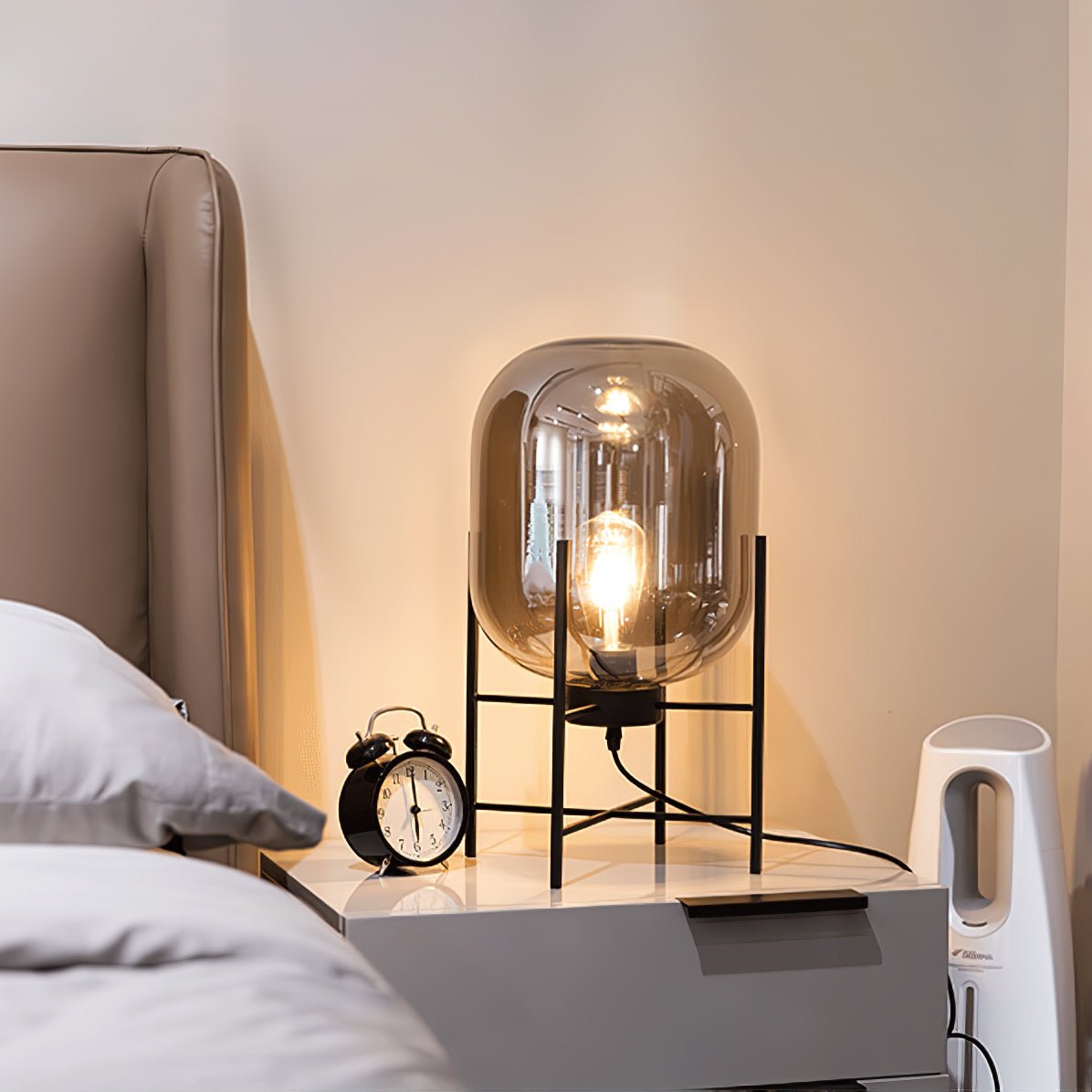 A dim, amber-lit glass shade on a black frame beside a bed with an alarm clock, from this Clérachic table lamp range.