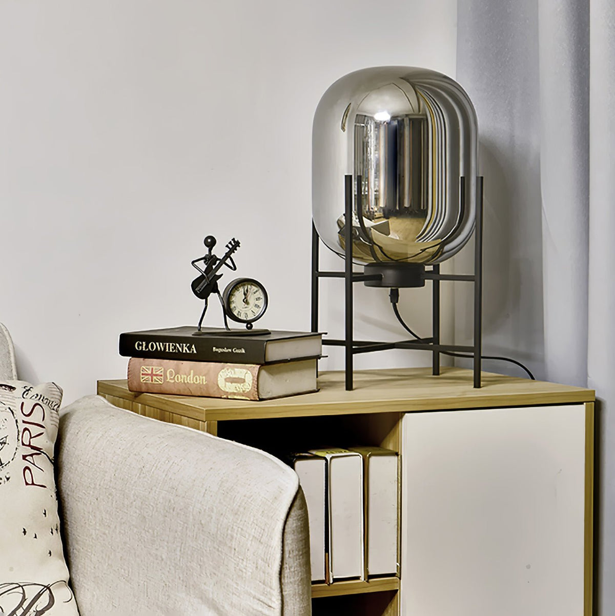 Mirrored smoke-grey glass shade on a black stand, this Clérachic lamp on a shelf beside a clock and a few books.