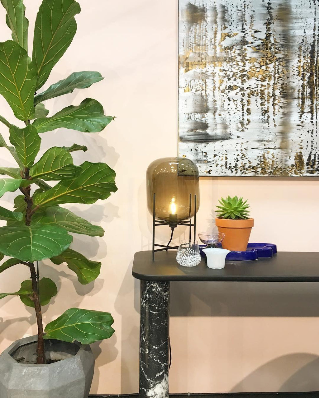 Amber glass lamp from Clérachic on a dark console table, beside a potted succulent and a tall green fiddle-leaf plant.
