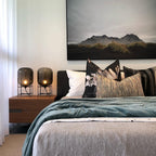 Two small glass table lamps from Clérachic lit on either side of a bed with a walnut bedside unit and a mountain print above.
