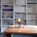 Clear smoky glass shade with a single filament bulb lit on a black frame, this Clérachic lamp on a timber desk before white shelves.
