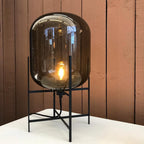 Dark smoked glass shade with a bright filament bulb inside, this Clérachic lamp on white paper against a rust-coloured wall.