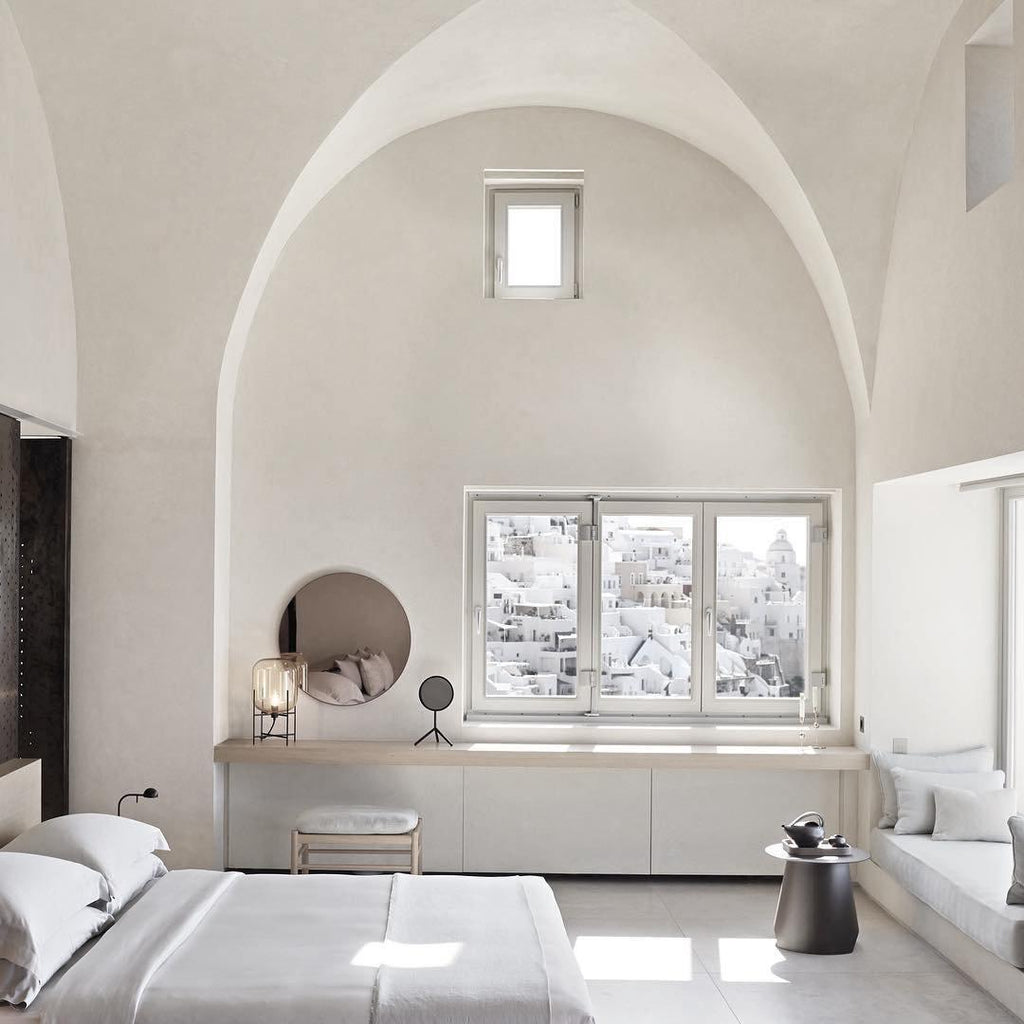 Tiny Clérachic table lamp on a low shelf in a pale vaulted bedroom with a window, a round mirror and a white bed.