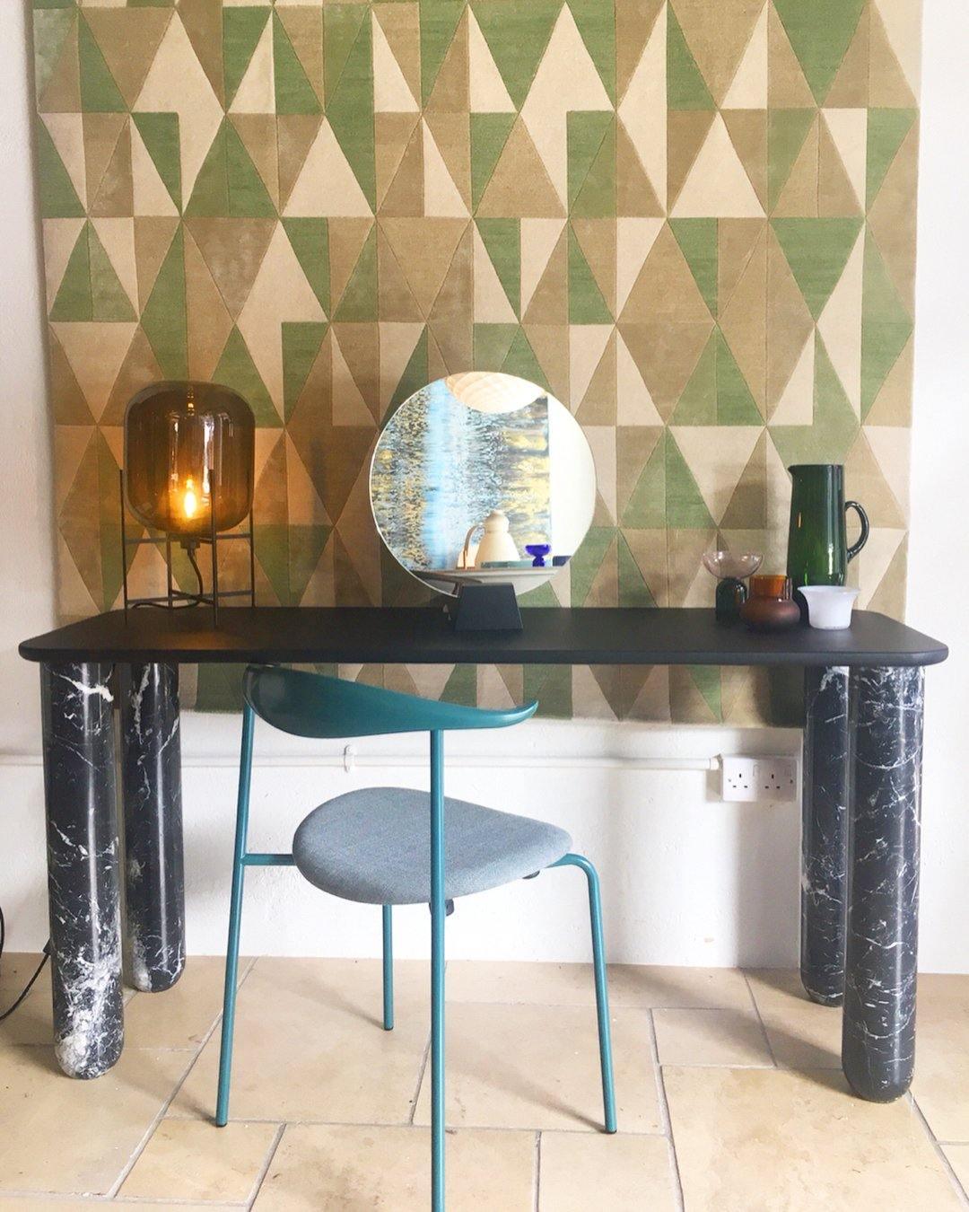 A dark amber glass lamp from Clérachic on a black marble-legged console, with a round mirror and a green geometric wall panel.