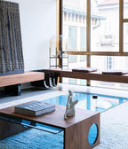 Slim Clérachic table lamp beside tall windows in a living space, with a walnut coffee table in the foreground.