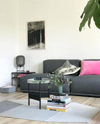 A smoked grey Clérachic lamp at the left of a dark grey sofa with pink cushions, in a bright room with a glass-topped coffee table.