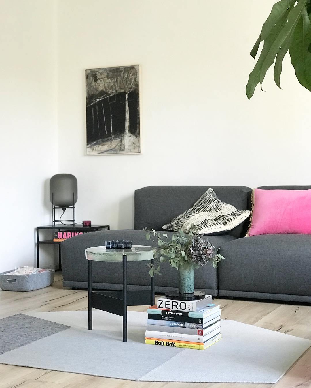 A smoked grey Clérachic lamp at the left of a dark grey sofa with pink cushions, in a bright room with a glass-topped coffee table.