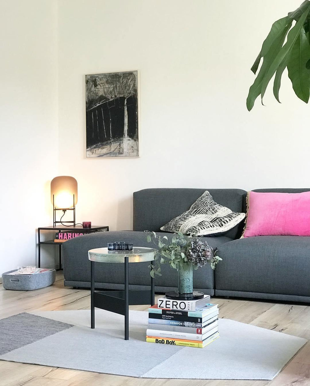 This Clérachic lamp glows softly in the corner of a living room, in front of a dark sofa with a bright pink cushion.