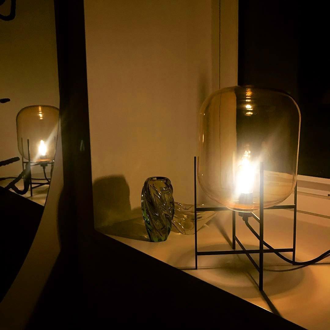 Warm amber glass shade lit from within on a dark ledge beside a small green glass vase, this Clérachic table lamp at night.