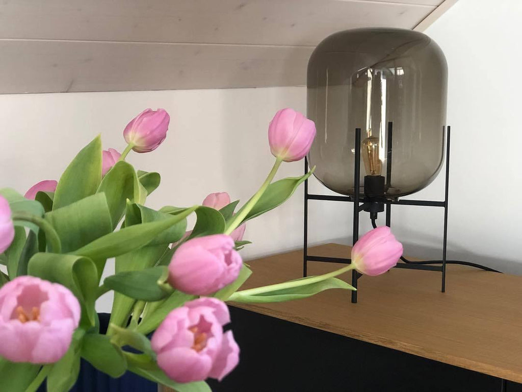 Grey-tinted glass shade on a black stand behind a bunch of pink tulips, this Clérachic table lamp on a wooden shelf.