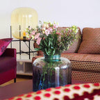 A pale yellow-amber glass shade on a black frame, this Clérachic lamp behind a vase of pink flowers in a colourful living room.