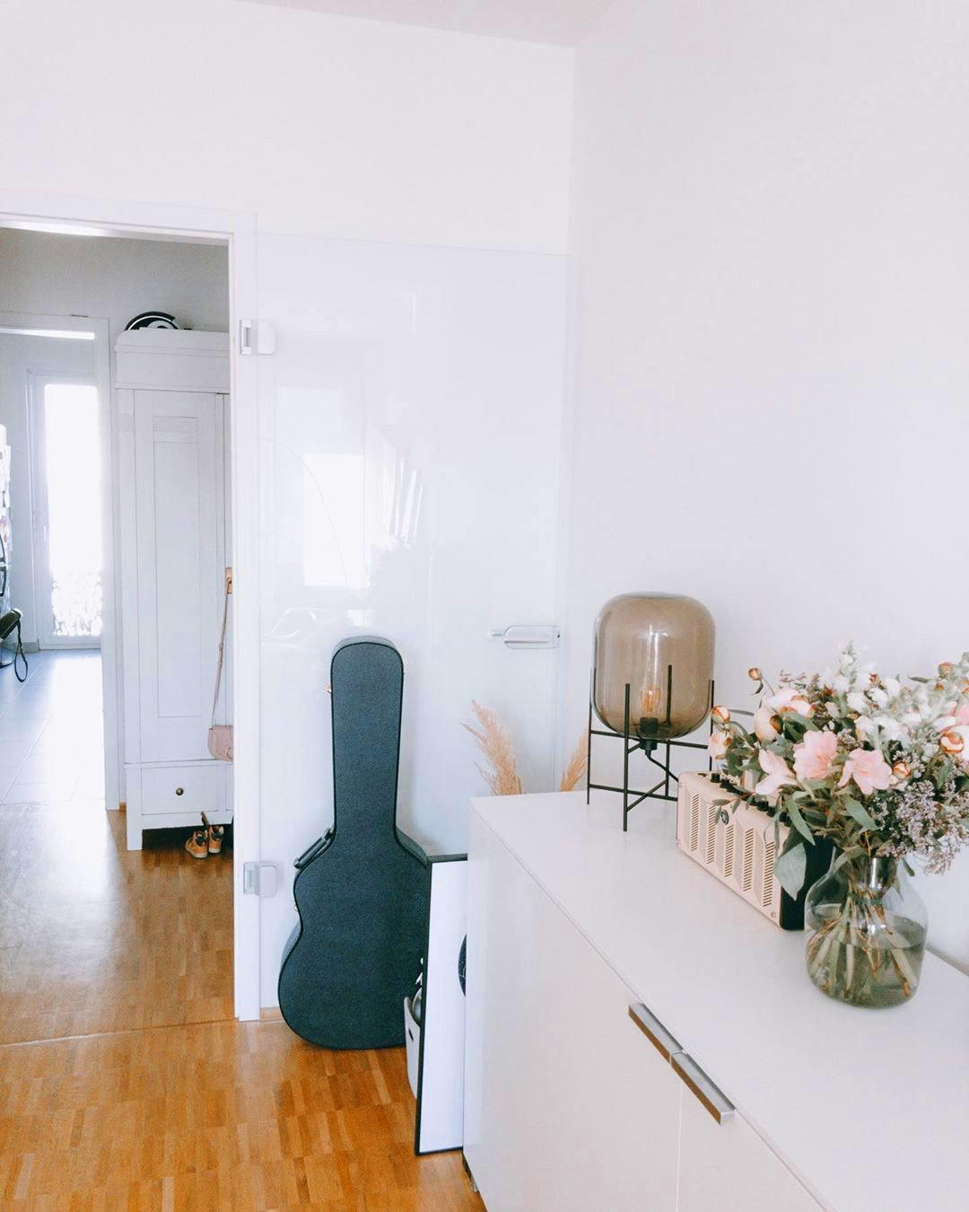 Taupe glass Clérachic table lamp on a white sideboard, beside a bouquet of pink blooms and a guitar case in a narrow hallway.