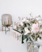 This Clérachic table lamp in smoky taupe glass stands beside a large bouquet of pale pink flowers on a white surface.