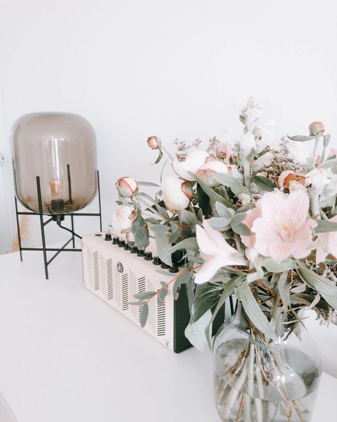 This Clérachic table lamp in smoky taupe glass stands beside a large bouquet of pale pink flowers on a white surface.