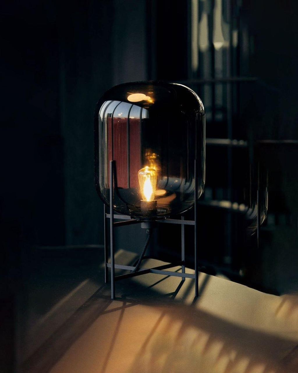 Dark smoked glass shade glowing on a black frame in a dim room, this Clérachic table lamp casting warm light on the floor.